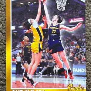 May include: Basketball trading card featuring Caitlin Clark, a point guard, in action. The card shows Clark in a yellow and black uniform, jumping to shoot a basketball. The card has a gold border and the text "Rookie F/X".