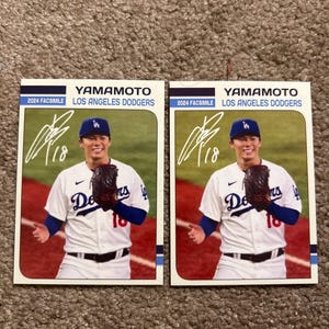 Peut inclure: Deux cartes de baseball avec un joueur en uniforme des Dodgers de Los Angeles. Les cartes affichent le nom "YAMAMOTO" et le texte "2024 FACSIMILE LOS ANGELES DODGERS". Le joueur porte une casquette bleue et tient un gant de baseball.