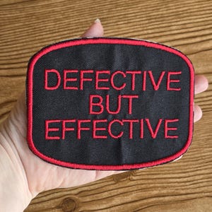 May include: Black rectangular patch with red border and lettering. The text "DEFECTIVE BUT EFFECTIVE" is embroidered in red thread. The patch is held in a hand against a wooden background.