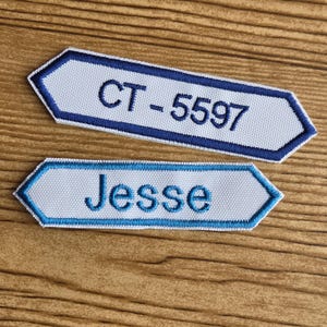 May include: Two embroidered name tags with a white background and blue trim. One tag reads "CT-5597" in blue, and the other reads "Jesse" in blue. The tags have a unique arrow-like shape.