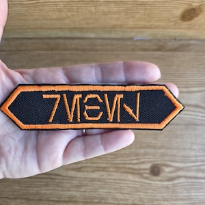 May include: Black and orange embroidered patch with the word "7IEVN". The patch has a black background with orange stitching around the edges and for the letters. The patch is held in a hand.
