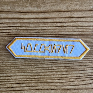 May include: A light blue embroidered patch with gold trim and text in a stylized font. The patch has a unique shape, with a light blue centre and gold border. The text is in a gold colour.