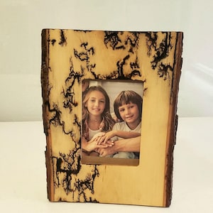 May include: A wooden picture frame with a natural bark edge and a light wood interior. The frame has a rectangular opening displaying a photo of two children. The wood has dark, intricate patterns.