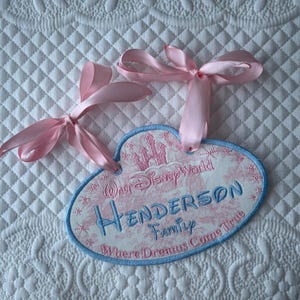 May include: An oval-shaped embroidered fabric plaque with a light blue border and two pink satin ribbons. The design features a pink castle, the text Walt Disney World, Henderson Family, and Where Dreams Come True in blue and pink embroidery.