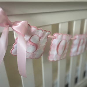 Puede incluir: Una pancarta de beb&eacute; de gingham rosa y blanco con la palabra "SO" bordada. La pancarta est&aacute; atada con una cinta rosa y cuelga de una cuna blanca.