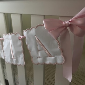 May include: A white fabric banner with scalloped edges and embroidered pink letters spelling "A-I-A-N". A large pink satin bow is tied to the crib. The banner is a decorative item for a nursery.