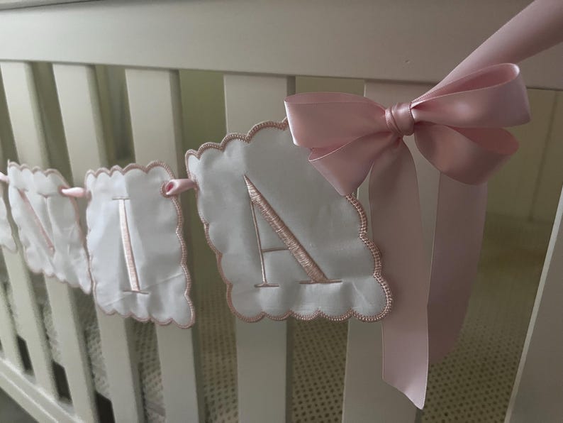 May include: A white fabric banner with scalloped edges and embroidered pink letters spelling "A-I-A-N". A large pink satin bow is tied to the crib. The banner is a decorative item for a nursery.