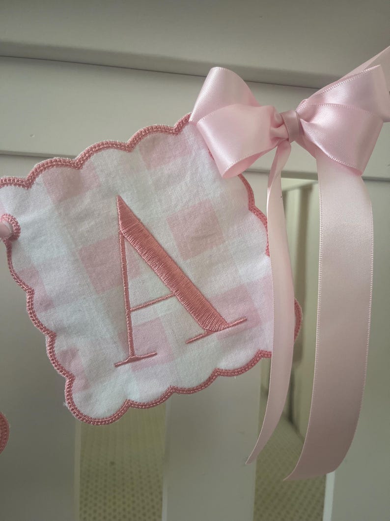 Puede incluir: Bandera de tela vichy rosa y blanco con la letra A bordada en hilo rosa. La bandera est&aacute; decorada con un lazo de sat&eacute;n rosa.