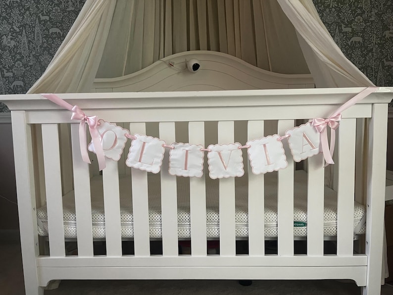 May include: A white crib with a canopy and a banner that spells "OLIVIA" in pink letters. The banner is made of white, scalloped-edged fabric squares with pink stitching and is strung with pink ribbon.