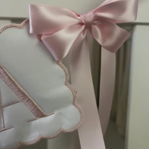 May include: A close-up of a baby nursery decoration. A white cloud-shaped banner with a pink embroidered letter "A" is attached to a white crib rail. A large, light pink satin bow with long streamers is tied to the top of the banner.