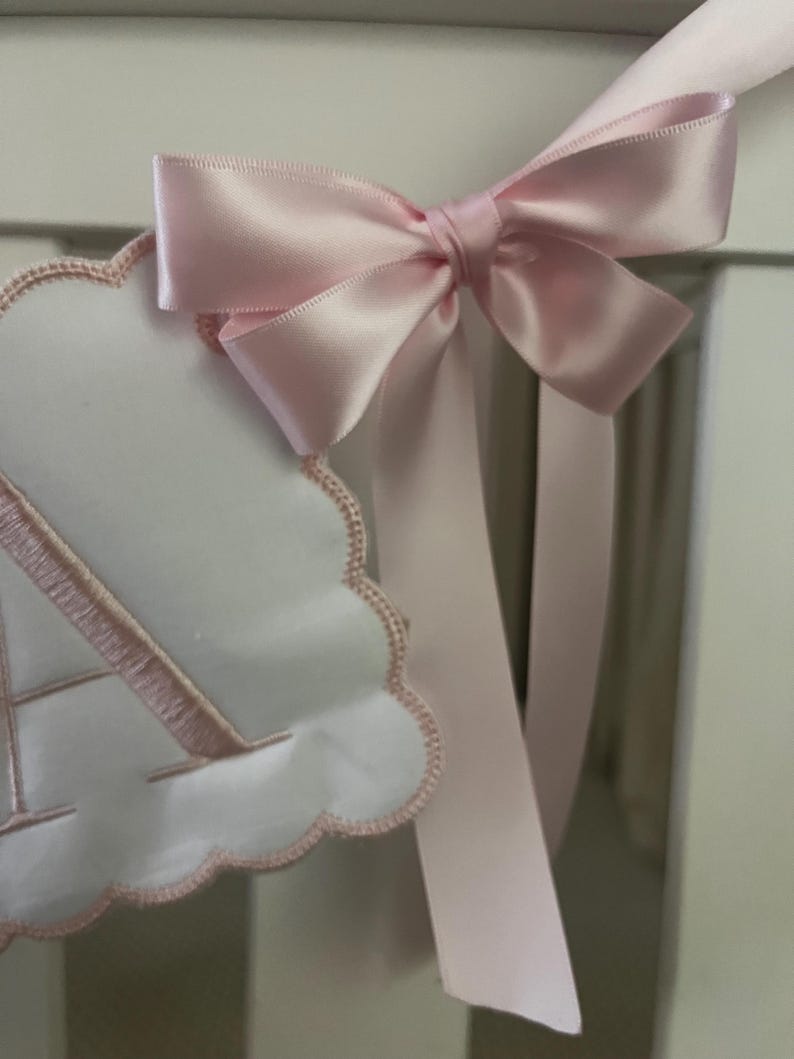 May include: A close-up of a baby nursery decoration. A white cloud-shaped banner with a pink embroidered letter "A" is attached to a white crib rail. A large, light pink satin bow with long streamers is tied to the top of the banner.