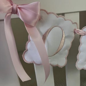 May include: A pink satin bow adorns a white banner with a scalloped edge. The banner features a stitched pink letter "O" and is part of a decorative nursery or baby room garland. The ribbon is a soft pink color.