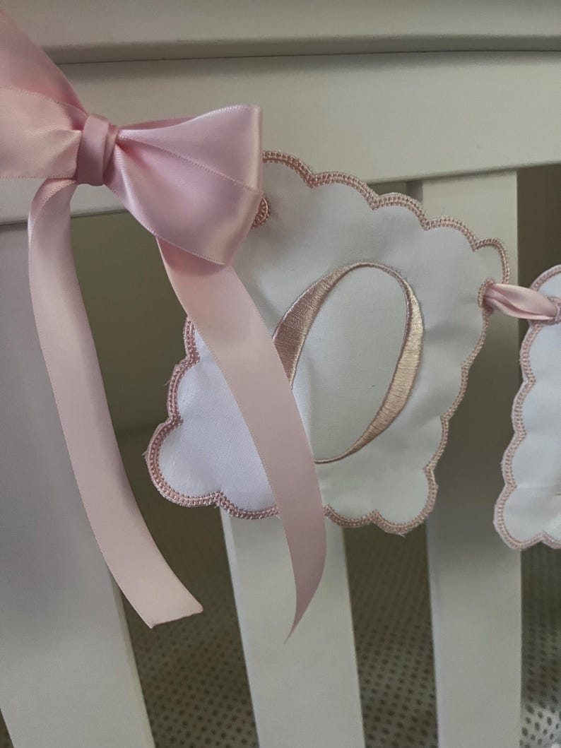May include: A pink satin bow adorns a white banner with a scalloped edge. The banner features a stitched pink letter "O" and is part of a decorative nursery or baby room garland. The ribbon is a soft pink color.