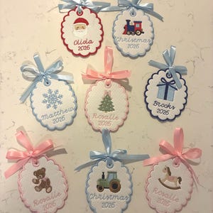 May include: Eight scalloped, embroidered fabric ornaments with satin ribbons. Designs include Santa, a train, a snowflake, a tree, a gift, a teddy bear, a tractor, and a rocking horse. Names and the year 2026 are stitched on each piece.