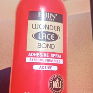 May include: Red spray bottle labelled "EBIN' NEW YORK WONDER LACE BOND ADHESIVE SPRAY EXTREME FIRM HOLD ACTIVE" with a gold circle sticker that says "BEAUTICIAN NO.1 CHOICE". The bottle is 2.7 fl.oz. (80ml) / NET WT. 60g e 2.1 oz.