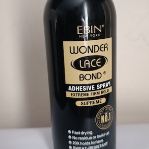 May include: A black can of Ebin Wonder Lace Bond adhesive spray. The can features gold lettering and a sticker with the words "Beautician's No. 1 Choice". The can is 2.78 oz (80 ml) and has a net weight of 60 g. The can is labelled as "Supreme" and "Extreme Firm Hold". The can has a clear plastic cap with a green button.