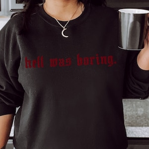 Op de afbeelding: Zwart sweatshirt met de rode tekst "hell was boring" in gotische letters. De top met lange mouwen heeft een ronde hals. Een persoon houdt een zwarte mok met een zilveren binnenkant vast.