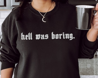 May include: A black sweatshirt with the text "hell was boring." printed in white on the front. The sweatshirt is being worn by a person in a kitchen setting.