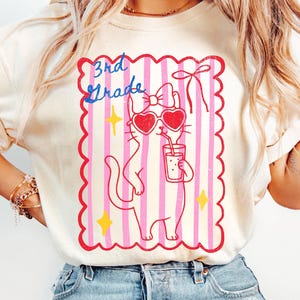 Puede incluir: Camiseta de color marfil con un gato de dibujos animados con gafas de sol en forma de corazón y bebiendo de un vaso. El gato está dentro de un marco de rayas rosas y blancas con la inscripción "3rd Grade" en azul.