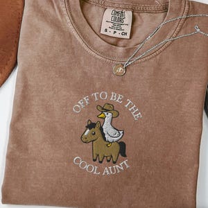 May include: Espresso-colored t-shirt with the embroidered phrase "Off to be the Cool Aunt" and a graphic of a horse and duck wearing a cowboy hat. A silver necklace with a pendant is draped across the shirt.