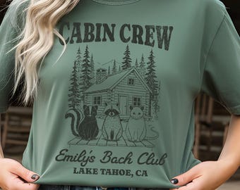 May include: Moss green t-shirt with a graphic that reads "CABIN CREW" above a cabin scene with a skunk, raccoon, and cat. The text "Emily's Bach Club, Lake Tahoe, CA" is below the graphic. The shirt is a relaxed fit.