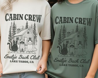 May include: Ivory and moss green t-shirts with the text "CABIN CREW" above a cabin illustration and the text "Emily's Bach Club LAKE TAHOE, CA". The ivory shirt is paired with blue shorts, and the moss shirt is paired with black shorts.