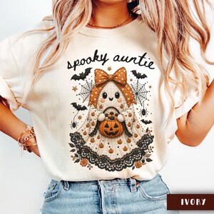 May include: An ivory t-shirt featuring a graphic of a ghost wearing a large orange bow, holding a jack-o'-lantern bucket. The design includes black lace trim, spiderwebs, bats, and small pumpkins, with the text "spooky auntie" printed above.