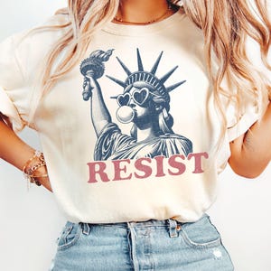 May include: Cream-colored t-shirt featuring a graphic of the Statue of Liberty wearing heart-shaped sunglasses and blowing a bubble. The word "RESIST" is printed in red below the image. The shirt is paired with blue jeans.
