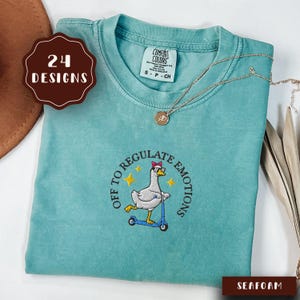 May include: A seafoam green crewneck t-shirt featuring an embroidered graphic of a goose wearing sunglasses and a pink bow, riding a blue scooter. The text reads OFF TO REGULATE EMOTIONS. The shirt has a Comfort Colors label and a thin gold necklace.