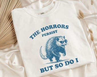 May include: White t-shirt with a blue graphic of an opossum wearing sunglasses, with the text "THE HORRORS PERSIST BUT SO DO I". The shirt is made of a soft material and is suitable for casual wear.