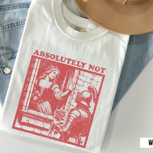 以下が含まれることがあります： 中世のシーンと「ABSOLUTELY NOT」のテキストが特徴の赤いグラフィックがプリントされた白いTシャツ。Tシャツは、青いジーンズとベージュの帽子の上に置かれています。