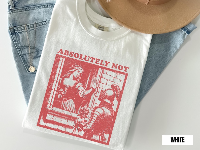 以下が含まれることがあります： 中世のシーンと「ABSOLUTELY NOT」のテキストが特徴の赤いグラフィックがプリントされた白いTシャツ。Tシャツは、青いジーンズとベージュの帽子の上に置かれています。