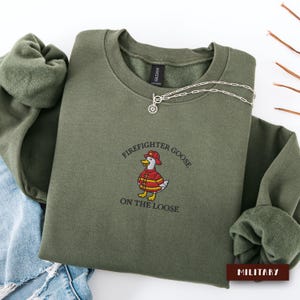 May include: An olive green sweatshirt with the text "Firefighter Goose On The Loose" and an embroidered cartoon goose in a firefighter uniform. A silver necklace with a sun pendant is draped on the sweatshirt. The word "MILITARY" is on a brown rectangle.