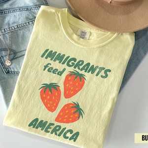Puede incluir: Una camiseta amarilla clara con el texto "IMMIGRANTS feed AMERICA" en verde, junto con tres fresas naranjas. La camiseta está colocada sobre unos vaqueros azules y un sombrero beige.