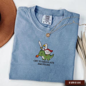May include: A blue Comfort Colors t-shirt featuring an embroidered design of a duck riding a green dragon, holding a sword. The text "OFF TO REGULATE EMOTIONS" is below the design. A gold necklace is draped across the shirt.