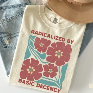 Könnte beinhalten: Elfenbeinfarbenes T-Shirt mit dem Text "RADICALIZED BY BASIC DECENCY" und einem floralen Design in Rot und Türkis. Das Shirt liegt auf einer blauen Jeans und einem beigen Hut.