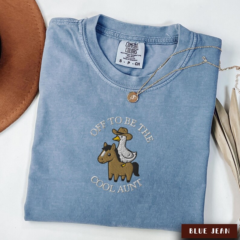 May include: Blue jean colored t-shirt with the embroidered phrase "Off to be the Cool Aunt". The design features a horse with a duck wearing a cowboy hat. A gold necklace and a brown hat are also visible.
