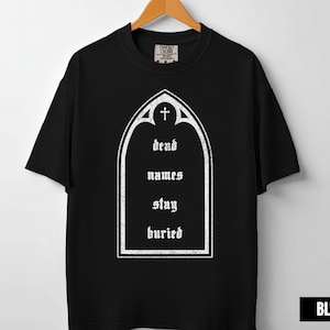 May include: Black t-shirt with a white graphic of a tombstone shape, a cross at the top, and the words "dead names stay buried" in a gothic font. The shirt is hanging on a wooden hanger.