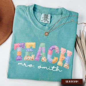 Könnte beinhalten: Türkisfarbenes T-Shirt mit dem Wort "TEACH" in bunten Patchwork-Buchstaben. Der Name "mrs. smith" steht darunter in Schreibschrift. Eine goldene Halskette mit Anhänger liegt auf dem Shirt.
