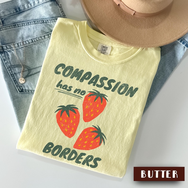 May include: A light yellow t-shirt with the words "COMPASSION has no BORDERS" in green, along with three red strawberries. The shirt is paired with blue jeans and a tan hat.