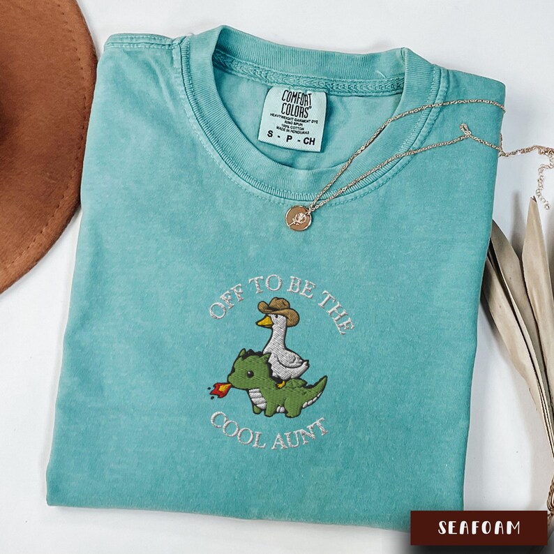 May include: Seafoam green t-shirt with the phrase "Off to be the Cool Aunt" embroidered in white thread. The design features a duck wearing a cowboy hat riding a green dinosaur. A gold necklace rests on the shirt.