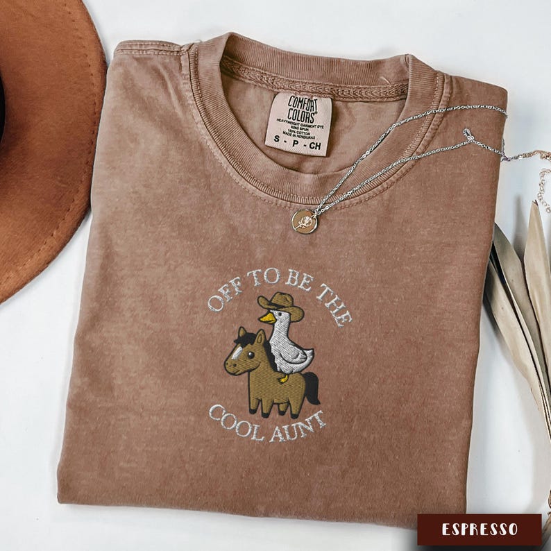 May include: Espresso-colored t-shirt with the embroidered phrase "Off to be the Cool Aunt" and a graphic of a horse and duck wearing a cowboy hat. A silver necklace with a pendant is draped across the shirt.