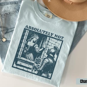 以下が含まれることがあります： 「絶対にダメ」の文字がプリントされた水色のTシャツ。その下に、女性と騎士の濃い青色のグラフィックがあります。女性は窓の中にいて、騎士は花を差し出しています。背景には、タン色の帽子と青いジーンズがあります。