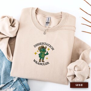 Puede incluir: Sudadera color arena con el texto "Dissertation Wrangler" y una rana de dibujos animados con sombrero de vaquero. La rana es verde con botas marrones y hace una señal de aprobación. Un delicado collar plateado descansa sobre la sudadera.