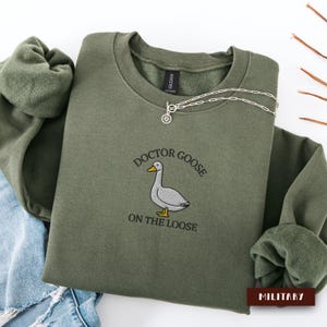 Puede incluir: Una sudadera verde oliva con la frase bordada "DOCTOR GOOSE ON THE LOOSE" y un dibujo de un ganso. Un collar plateado con un colgante de sol está drapeado sobre la sudadera. La palabra "MILITARY" está impresa en un rectángulo marrón.