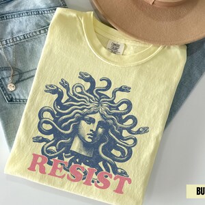Könnte beinhalten: Hellgelbes T-Shirt mit blauem Medusa-Motiv und dem rosa Wort "RESIST". Das Shirt liegt gefaltet auf einer blauen Jeans und einem beigen Hut.