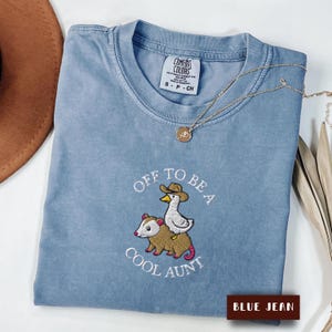May include: A blue jean coloured t-shirt featuring an embroidered graphic of a duck wearing a cowboy hat riding on the back of an opossum. The text "OFF TO BE A COOL AUNT" is embroidered in white around the graphic. The shirt has a Comfort Colors label.