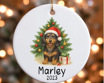 May include: A white, round Christmas ornament featuring a dachshund wearing a Santa hat in front of a Christmas tree. The ornament has the name "Marley" and the year "2023" printed on it. A gold string is attached for hanging.