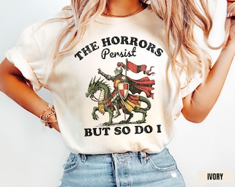 Può includere: T-shirt color avorio con grafica invecchiata di un cavaliere su un drago, con la scritta "THE HORRORS Persist BUT SO DO I". Il cavaliere è corazzato e porta uno scudo e una bandiera.