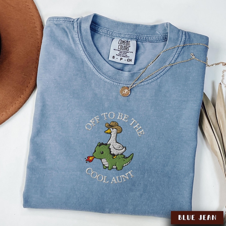 May include: A blue jean colored t-shirt with the embroidered phrase "Off to be the Cool Aunt". The design features a duck wearing a cowboy hat riding a green dinosaur. A gold necklace is draped across the shirt.